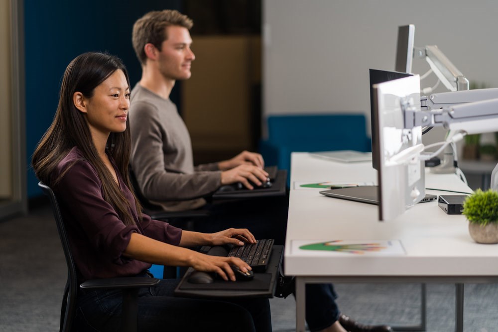 Two employees sitting a desk and using Kensington monitor arms to display their monitors