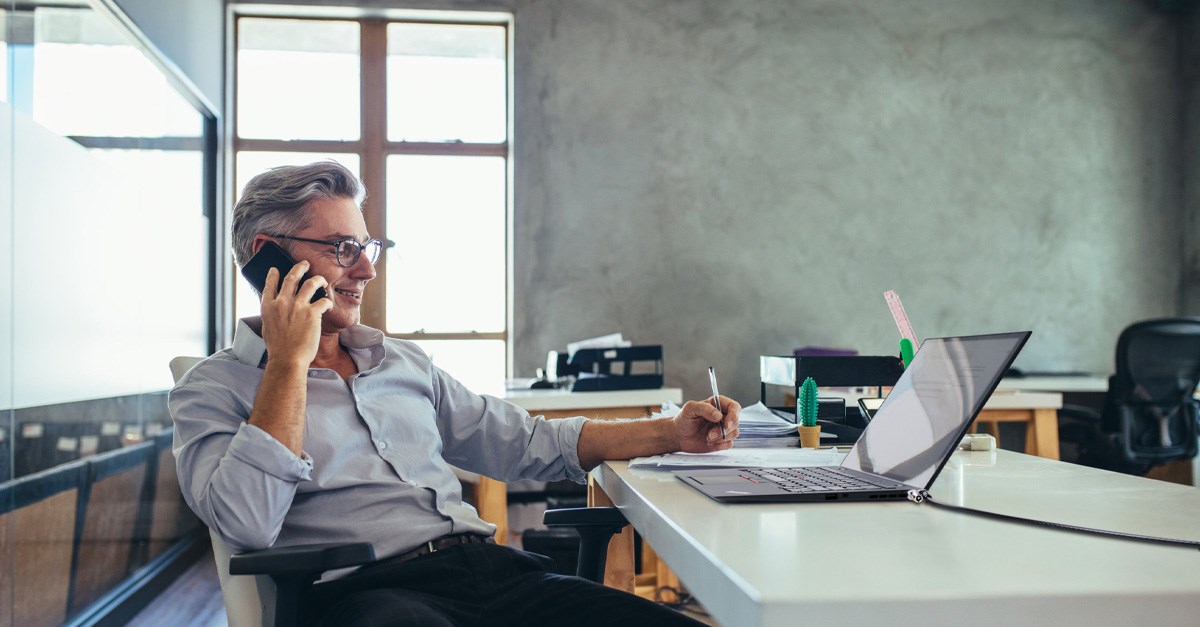 Man talking on a cell phone, taking notes, and sitting in front of a laptop