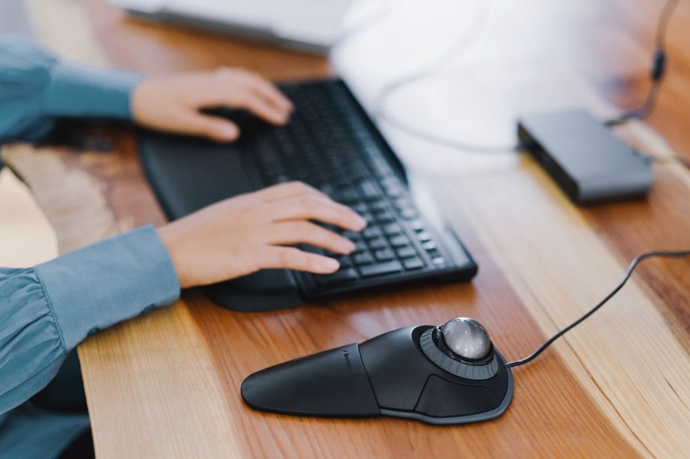 Person typing on a keyboard and using a Kensington Orbit Trackball Mouse with Scroll Ring 