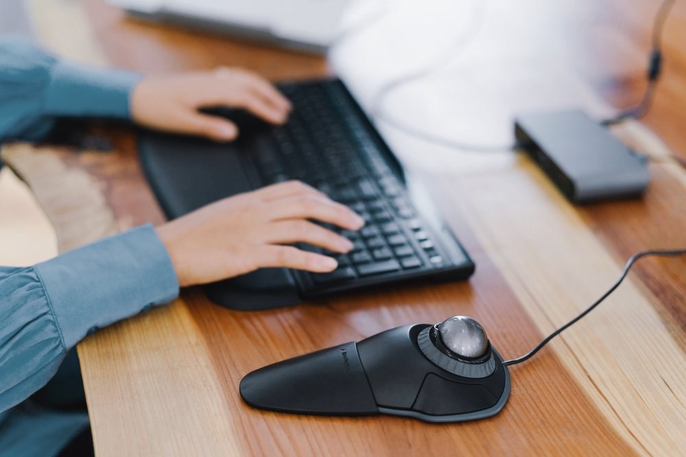 Person typing on a keyboard and using a Kensington Orbit Trackball Mouse with Scroll Ring