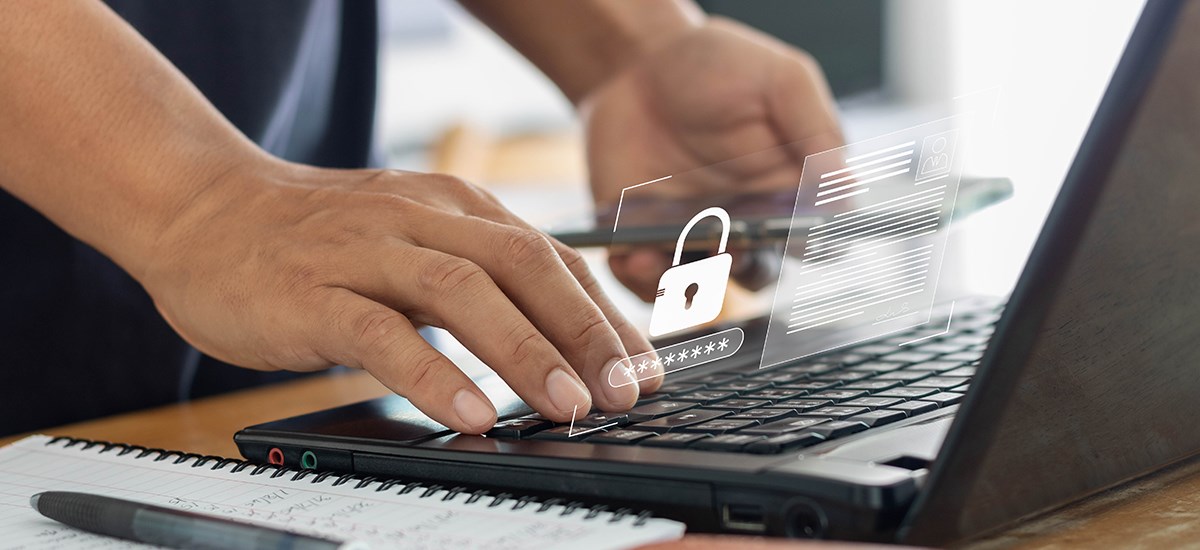 Hands typing a password on a laptop using a cellphone as password manager.