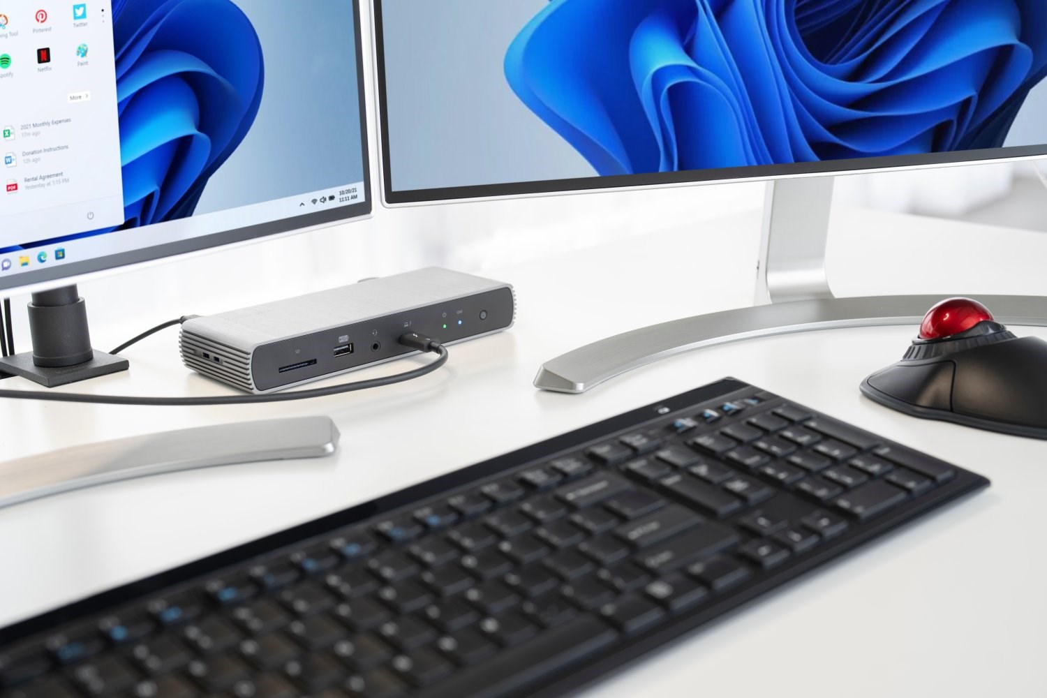 A close-up view of a docking station on a desk, with a keyboard, trackball, and two monitors displaying a Windows screensaver.