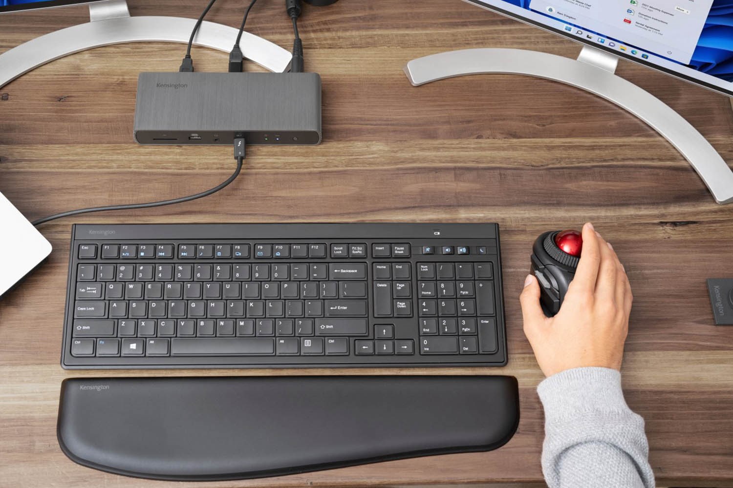 The SD5750T docking station connected to a monitor, with a keyboard, wrist rest, and a hand using a trackball mouse on a wooden desk.