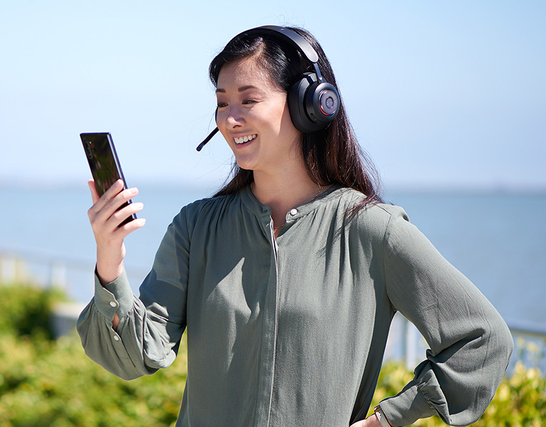 Woman stading outside with the ocean behind her looking at her phone and wearing a Kensington H3000 headset