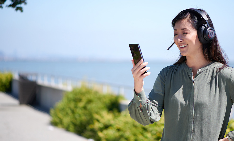 Woman stading outside with the ocean behind her looking at her phone and wearing a Kensington H3000 headset