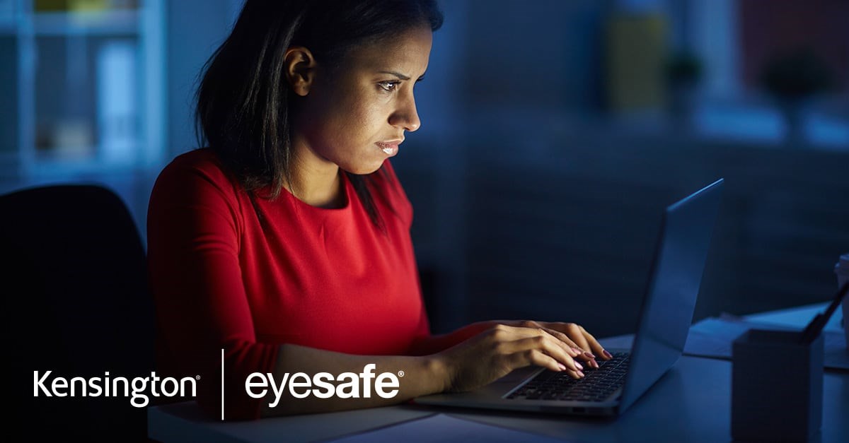 Professional woman in red shirt working on laptop in dimly lit office environment, with Kensington and Eyesafe logos displayed