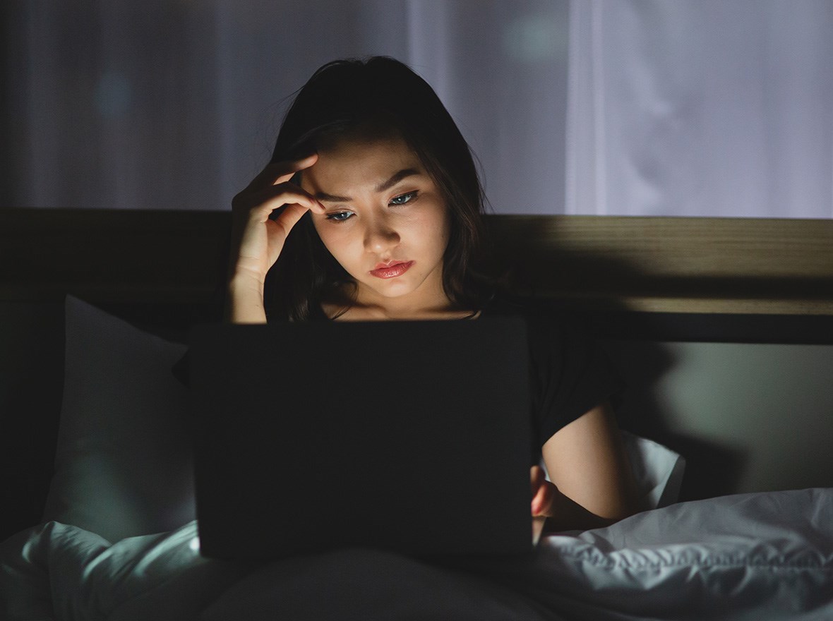 Woman sitting in dark room on bed using bright laptop.