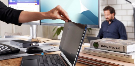 Woman placing privacy screen on laptop in office with man on laptop in background.