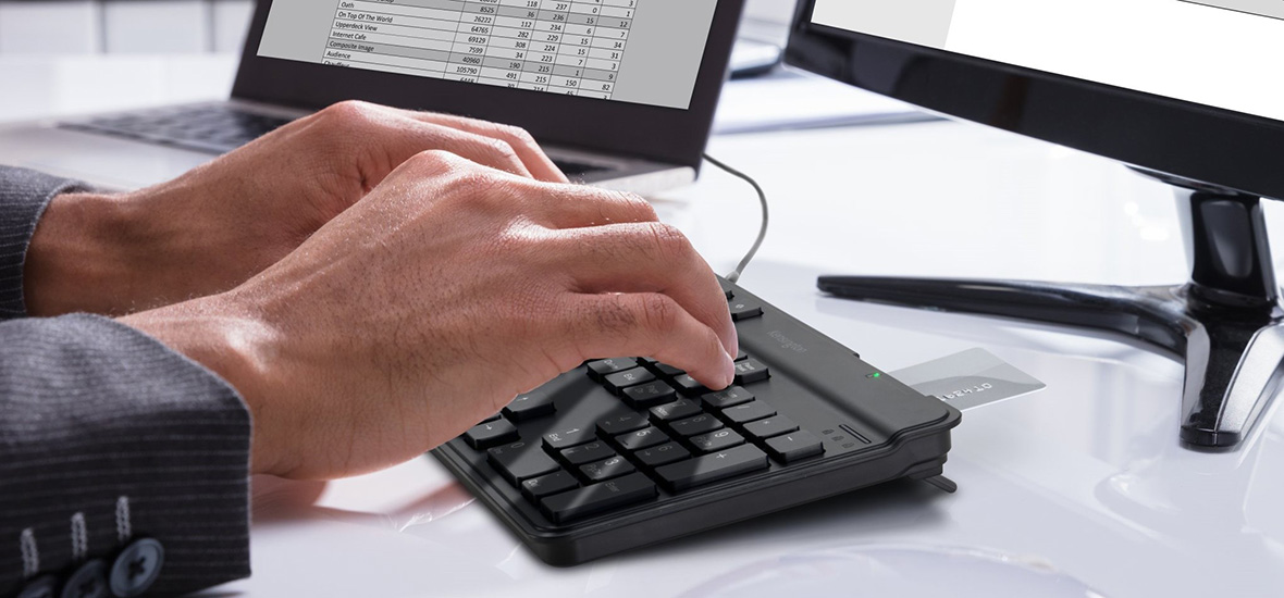 Hands typing on a keyboard with a smart card inserted, connected to a computer monitor and laptop